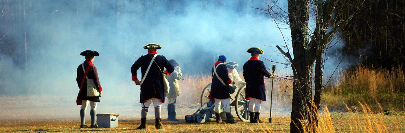 Men dressed in Revolutionary War uniforms firing a cannon in a field.