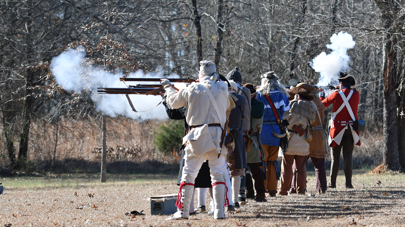 A group of Revolutionary War reenactors dressed in Revolutionary War clothing standing in a line firing rifles.