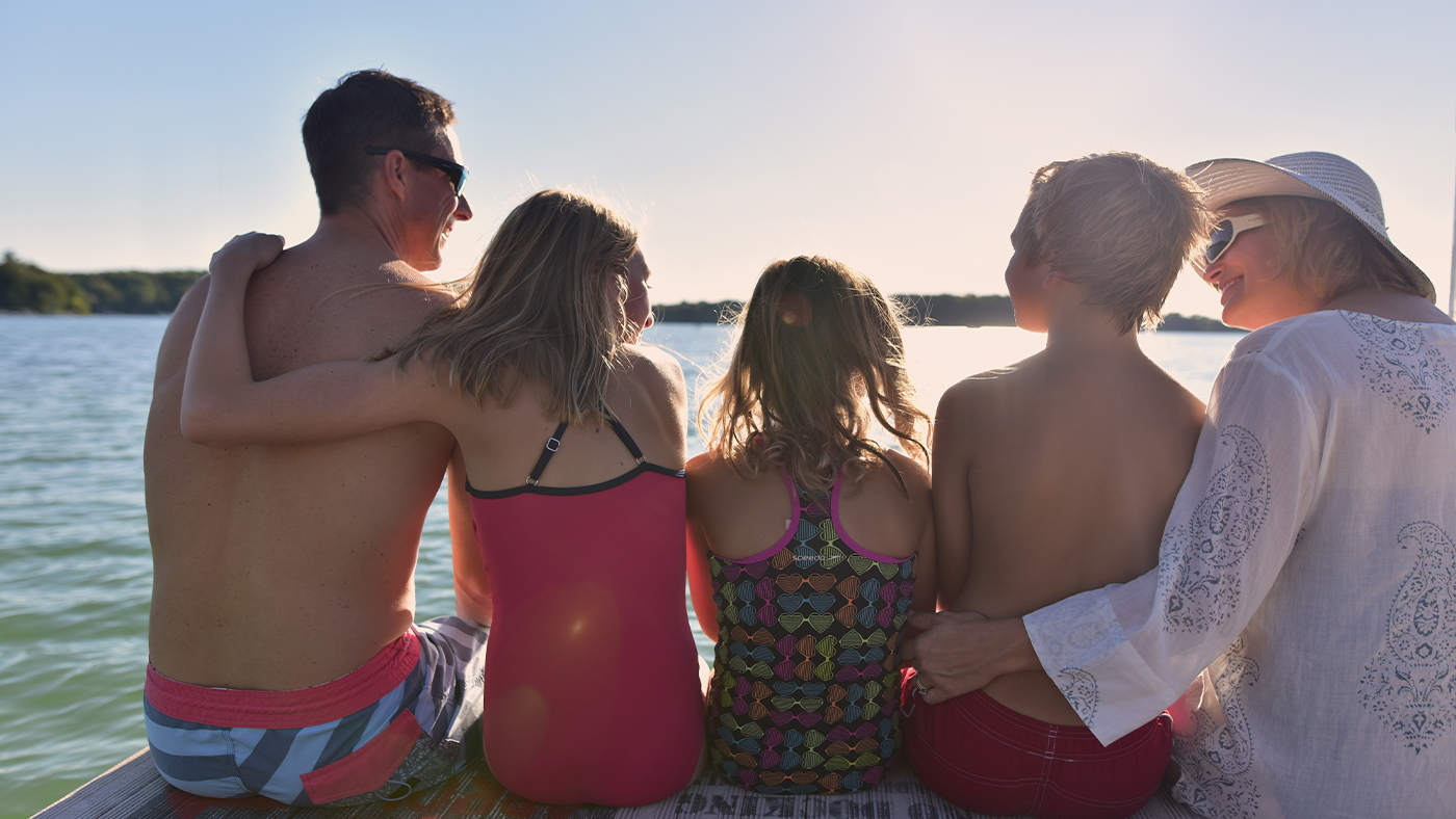 Mom, dad, two daughters and son wearing swim suits sitting on a dock looking out over the lake.