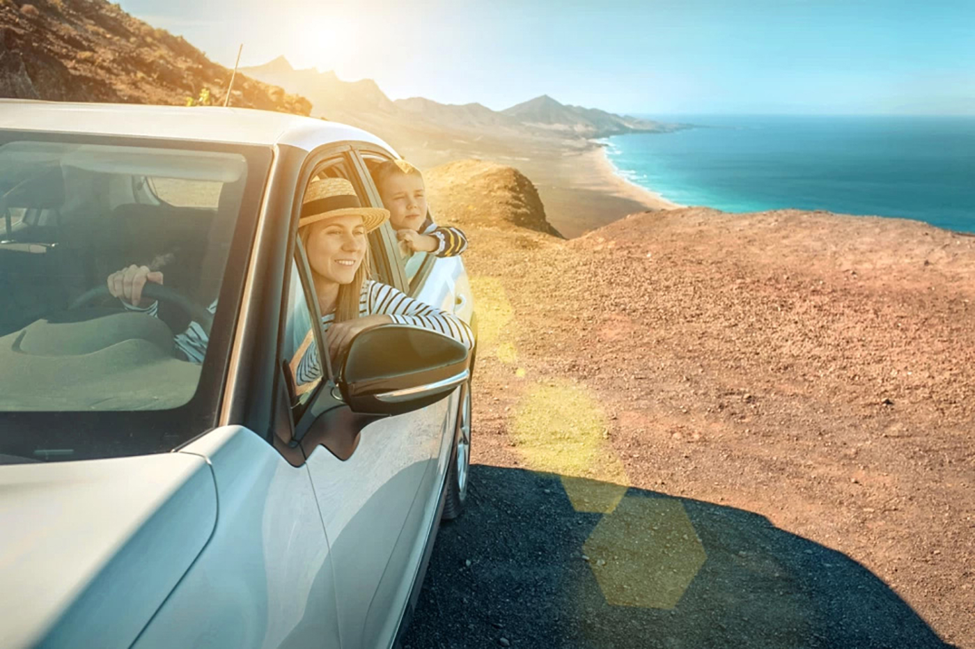 A woman wearing a straw hat and a striped shirt and a young girl wearing a striped shirt looking out the driver's side window of a white car parked in a landscape with hills and a body of water in the background.