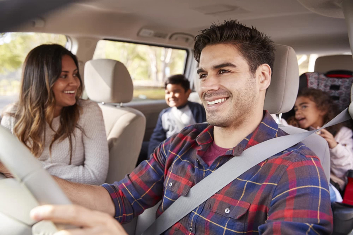 Dad with brown hair wearing a blue and red plaid shirt, mom wearing a white long-sleeve shirt in the front seat of a car wearing seatbelts and two kids in the back seat wearing seatbelts.