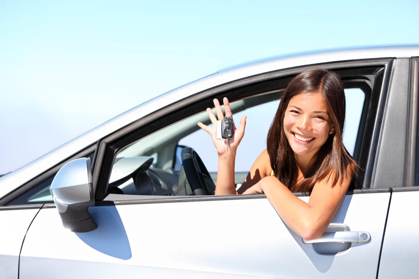 A young, smiling woman with brown hair looking out the driver's rolled down window of a white car with the car's key fob ring around one of the fingers of her right hand.