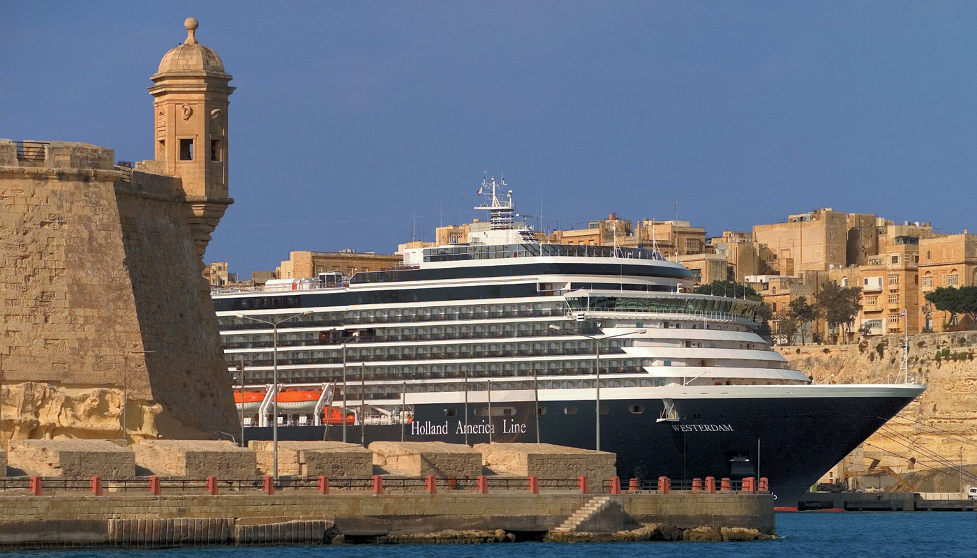 A large Holland America cruise ship docked in the medieval port on the island of Malta. There  are sand-colored brick buildings surrounding it and the water is a deep beautiful blue.