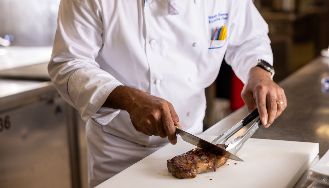 A Holland America chef prepares to cut a grilled steak. He is wearing the classic white chef's clothing.