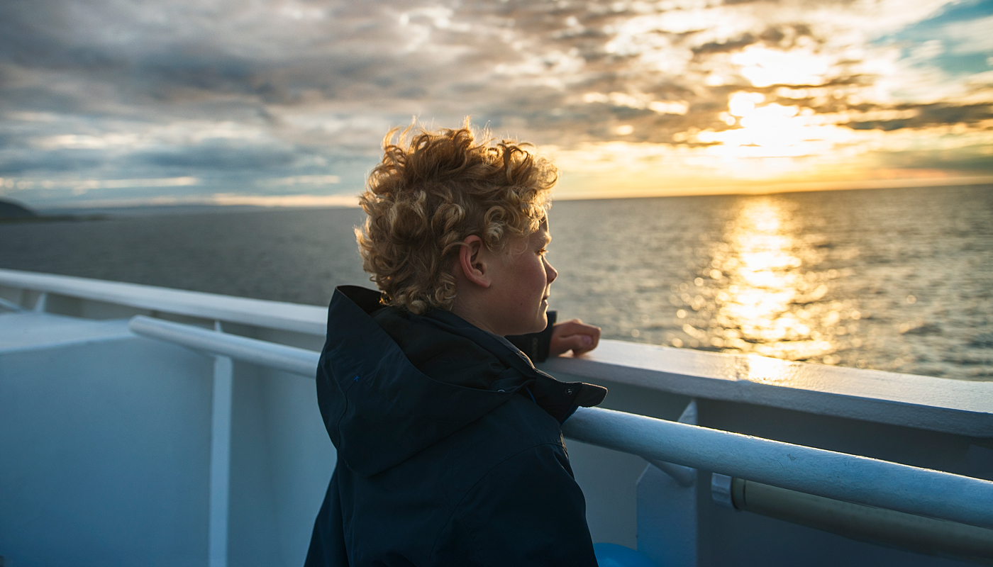 A young woman looking over the railing of a ship out to sea with a setting sun off in the distance.