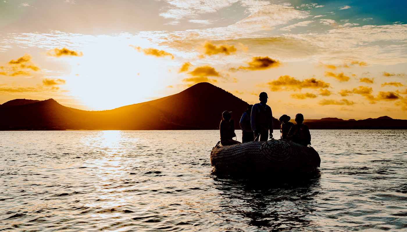 Sunsetting behind an island with a mountain and a boat in the foreground with about 5 people on board.