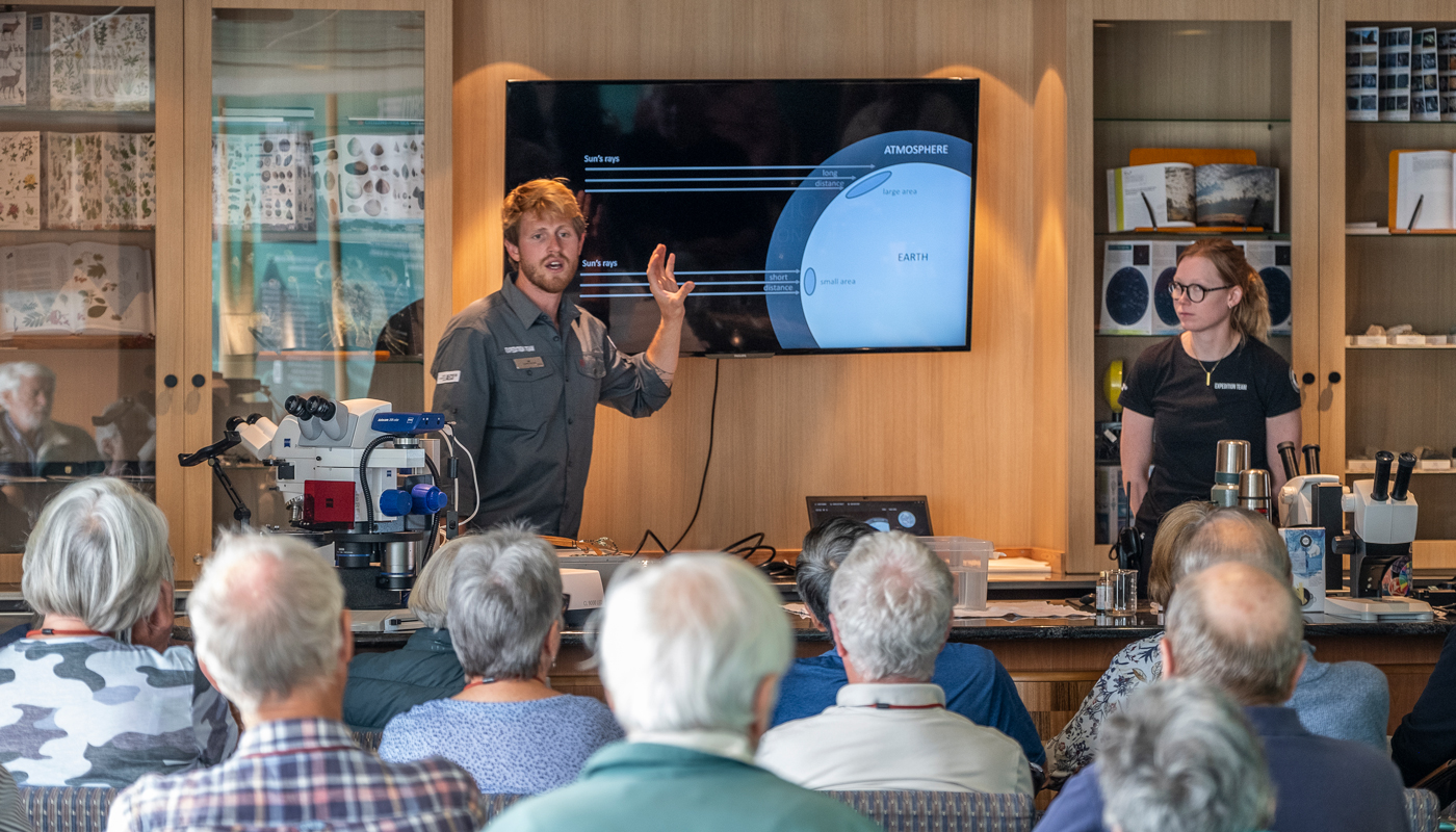 A scientist pointing to a screen with information about the local ecosphere with a dozen passengers looking on as he talks.