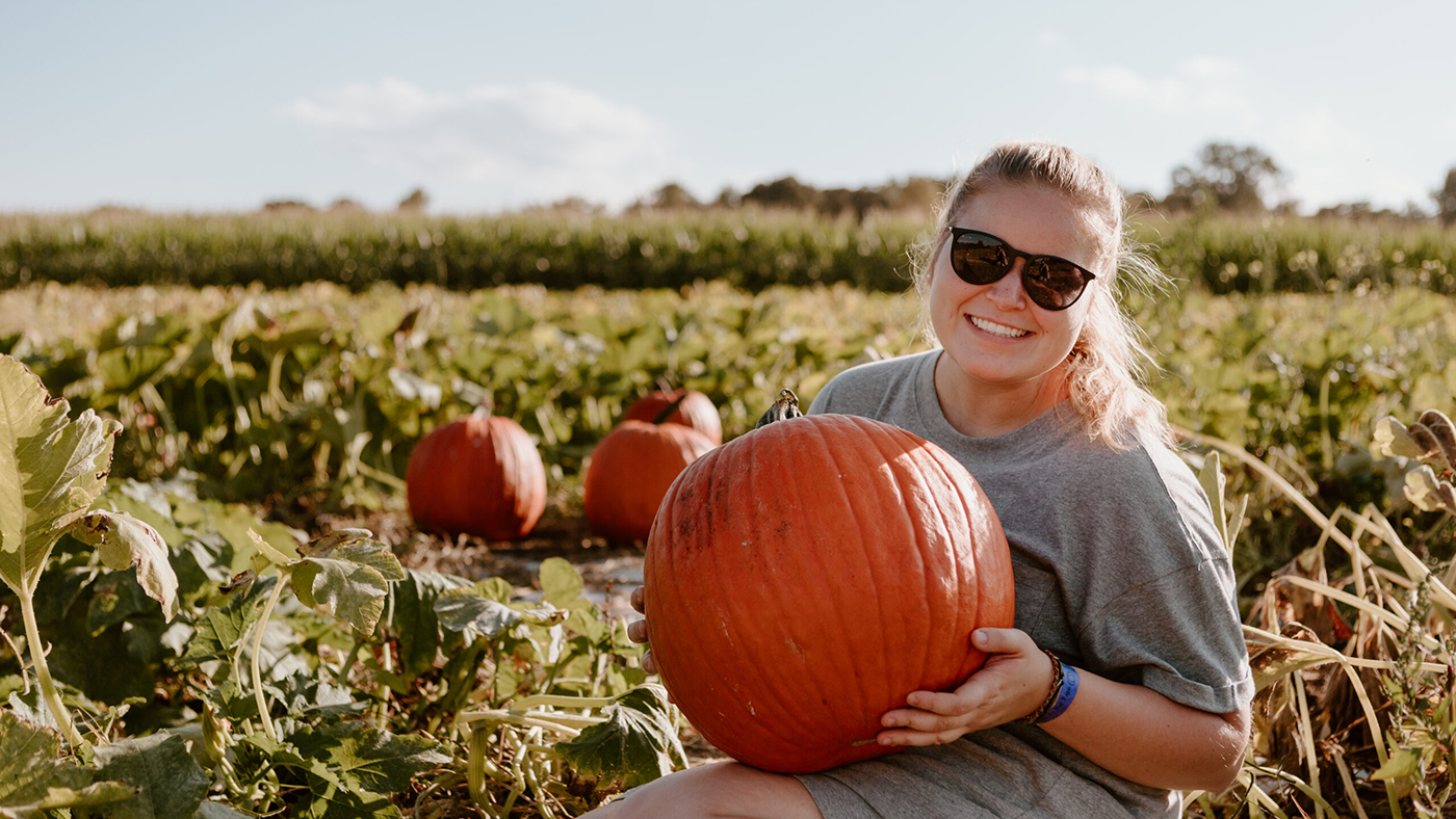 Woman wearing a gray t-shirt , shorts and sunglasses holding a large pumpkin in a pumpkin patch.