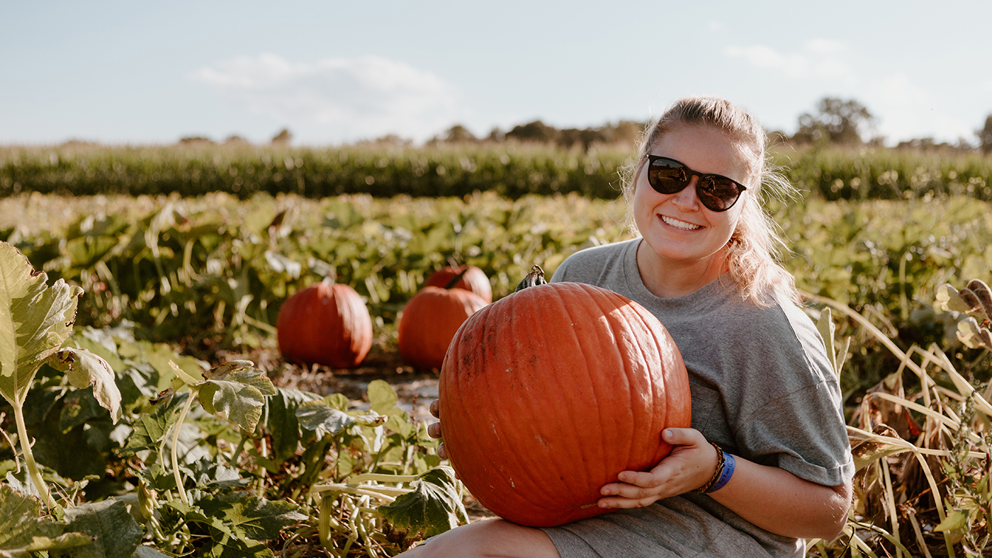 Smiling woman wearing sunglasses holding a pumpkin in a pumpkin patch.