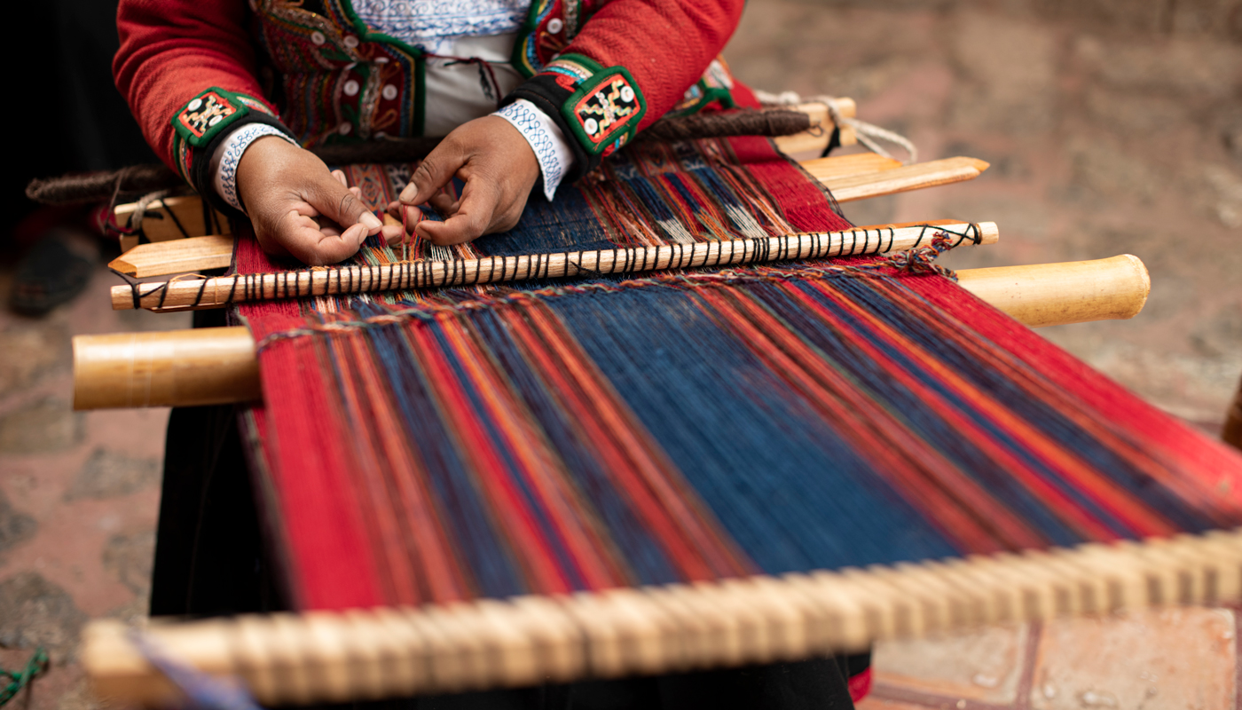 A indigenous person weaving a blanket of multi-colors on an old loom.